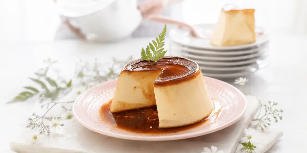Delicioso flan de queso con caramelo líquido, decorado con una hoja verde. En el fondo, una persona prepara más postres.