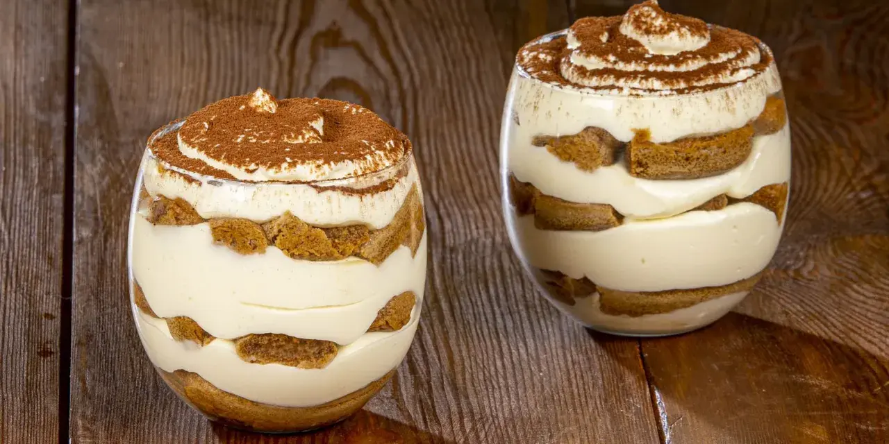 Due bicchieri di tiramisù senza uova, stratificati con crema e savoiardi, spolverati di cacao.