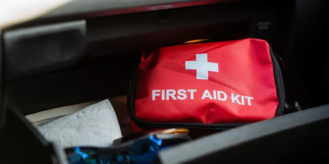 Czerwona apteczka samochodowa z białym krzyżem i napisem "FIRST AID KIT" leży w schowku. Obok niej papierowy ręcznik.