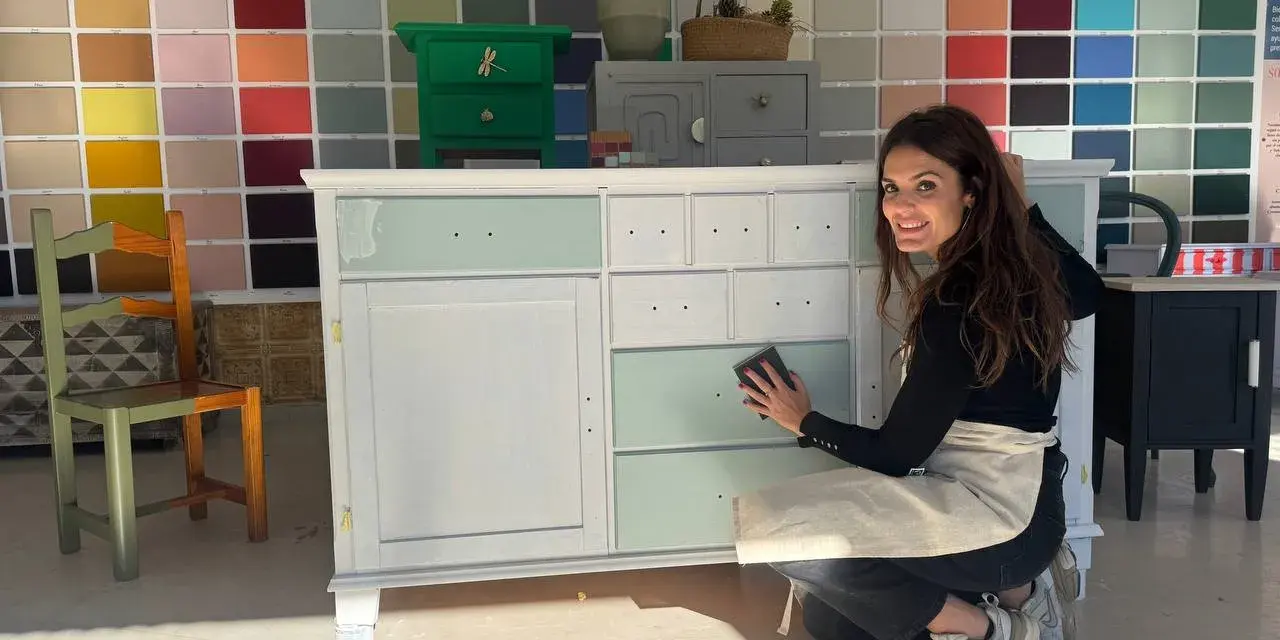 Mujer sonríe mientras lija un mueble para pintar. Detrás, un muro con muestras de colores de pintura.