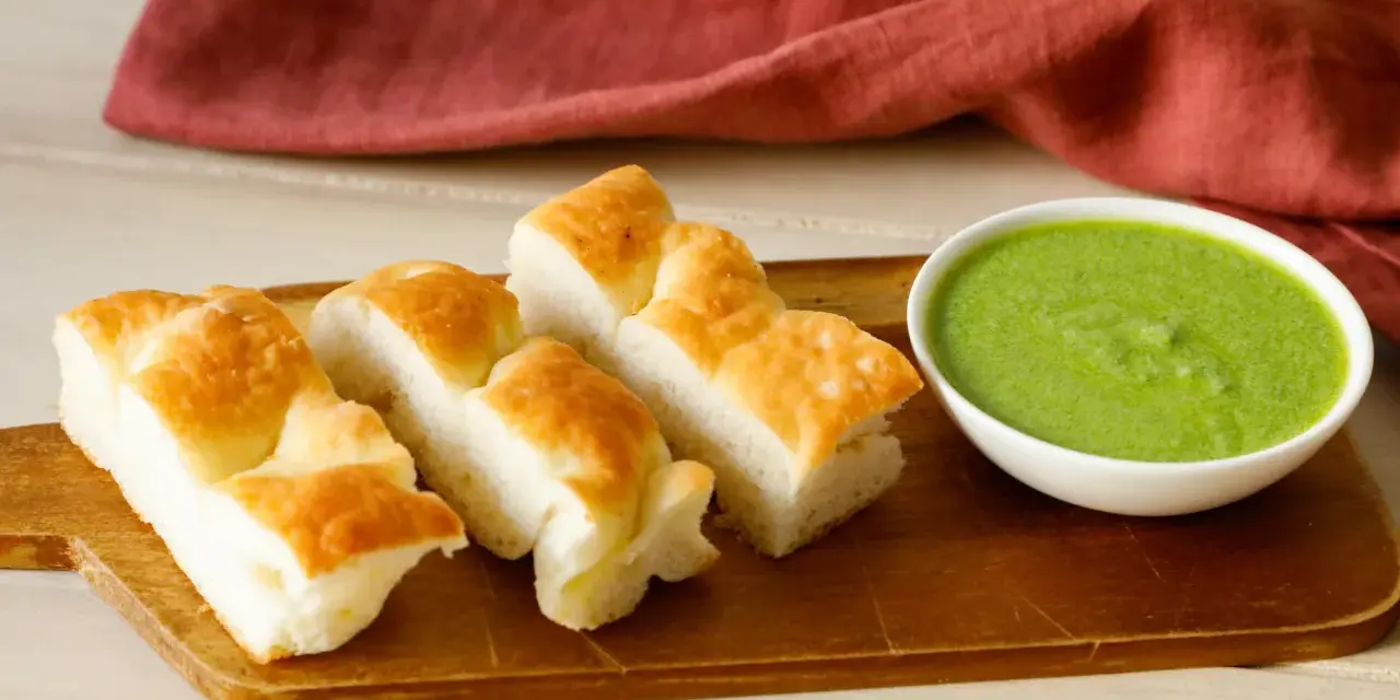 Focaccia tagliata a pezzi su tagliere di legno, accanto a una ciotola di pesto di salvia.