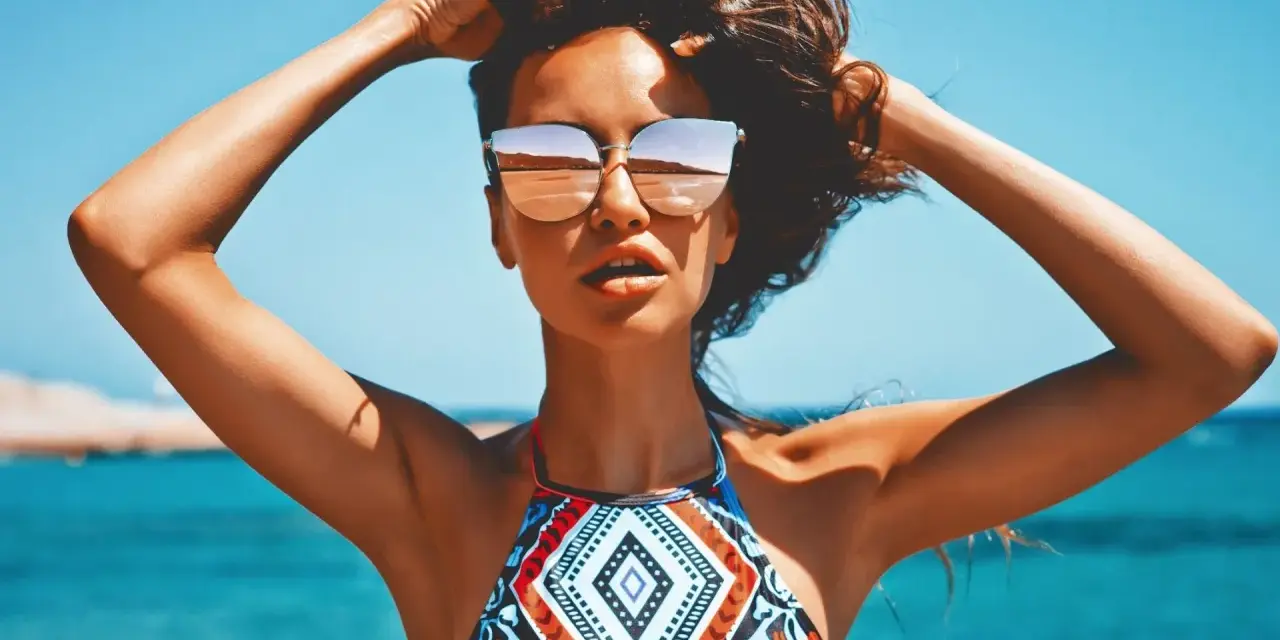 Kobieta w okularach przeciwsłonecznych na plaży, z rękami w gorze. Jej skóra ma piękny, złocisty odcień, jakby świadczył o wspaniałych beta karoten efektach.