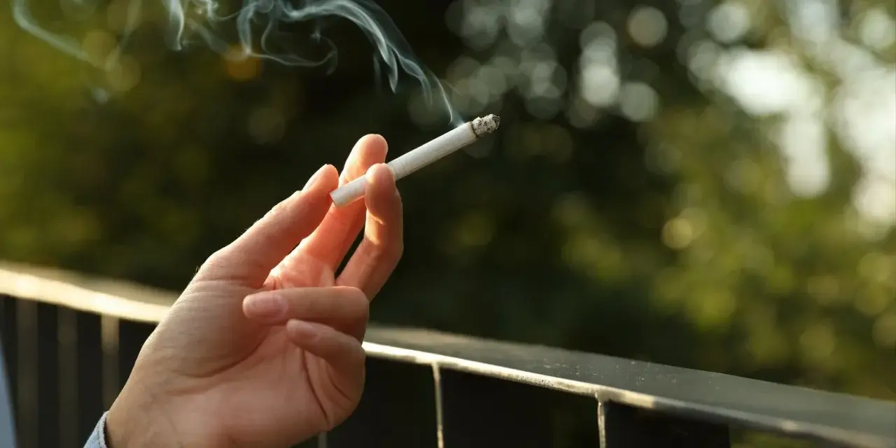 Dłoń trzyma zapalonego papierosa, z którego unosi się dym. Czy to prawda o rzekomym zakazie palenia papierosów na balkonach w nowych regulaminach?