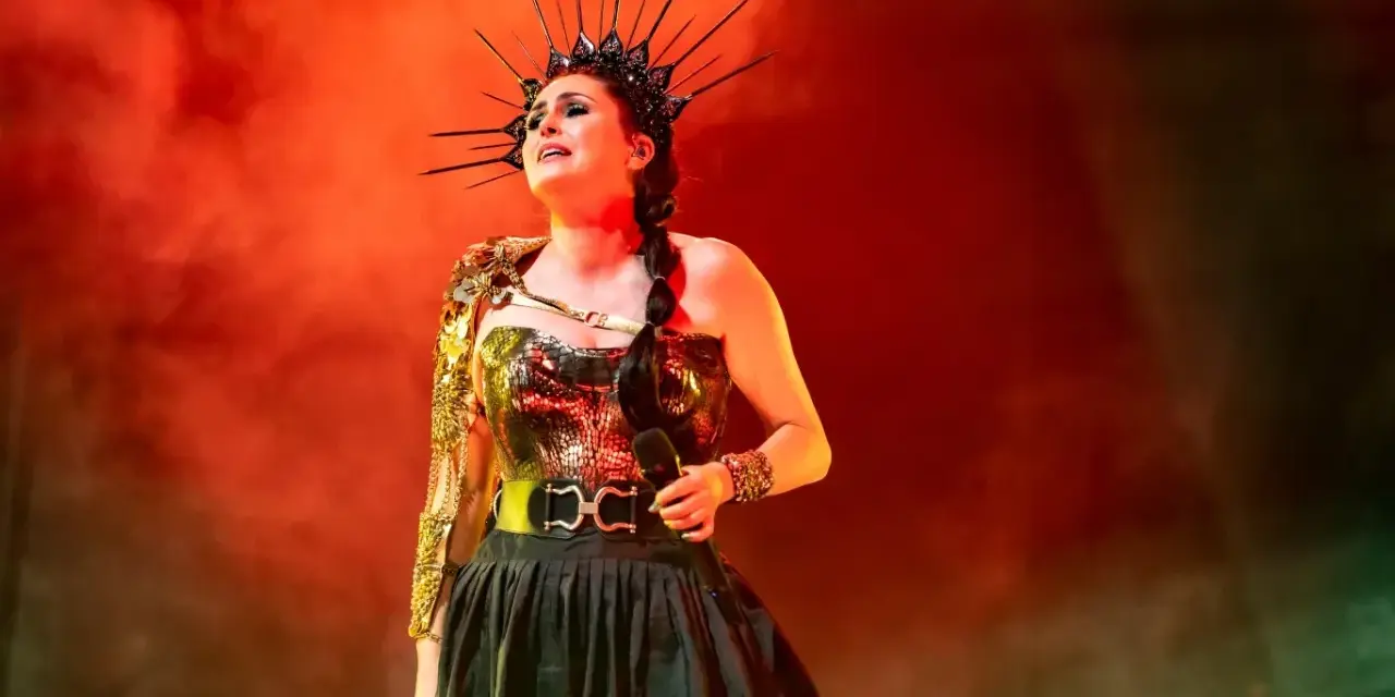 Wokalistka Within Temptation w koronie i błyszczącej kreacji, z mikrofonem w dłoni, na tle czerwonej mgły.