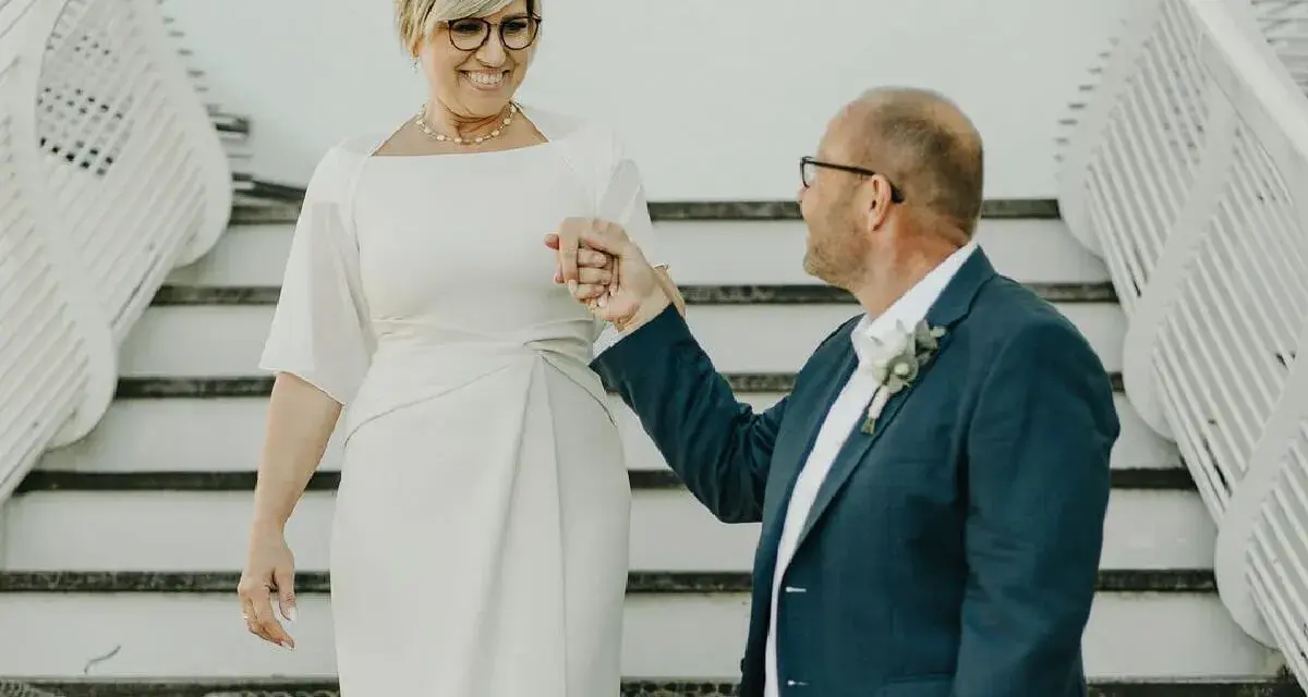 Elegancka kobieta po pięćdziesiątce w sukience do ślubu cywilnego, trzymająca za rękę pana młodego w granatowym garniturze.