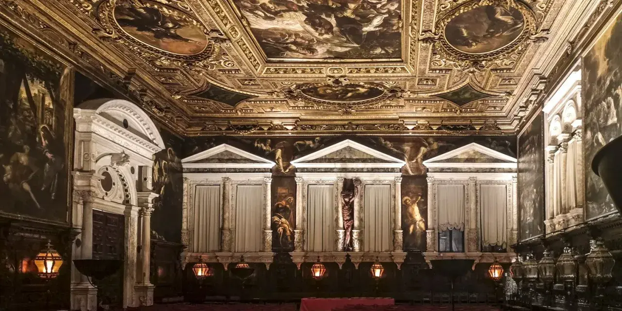 Sala capitolare Scuola Grande di San Rocco: soffitto dorato affrescato, pareti con dipinti e statue, sedie rosse disposte per un evento.