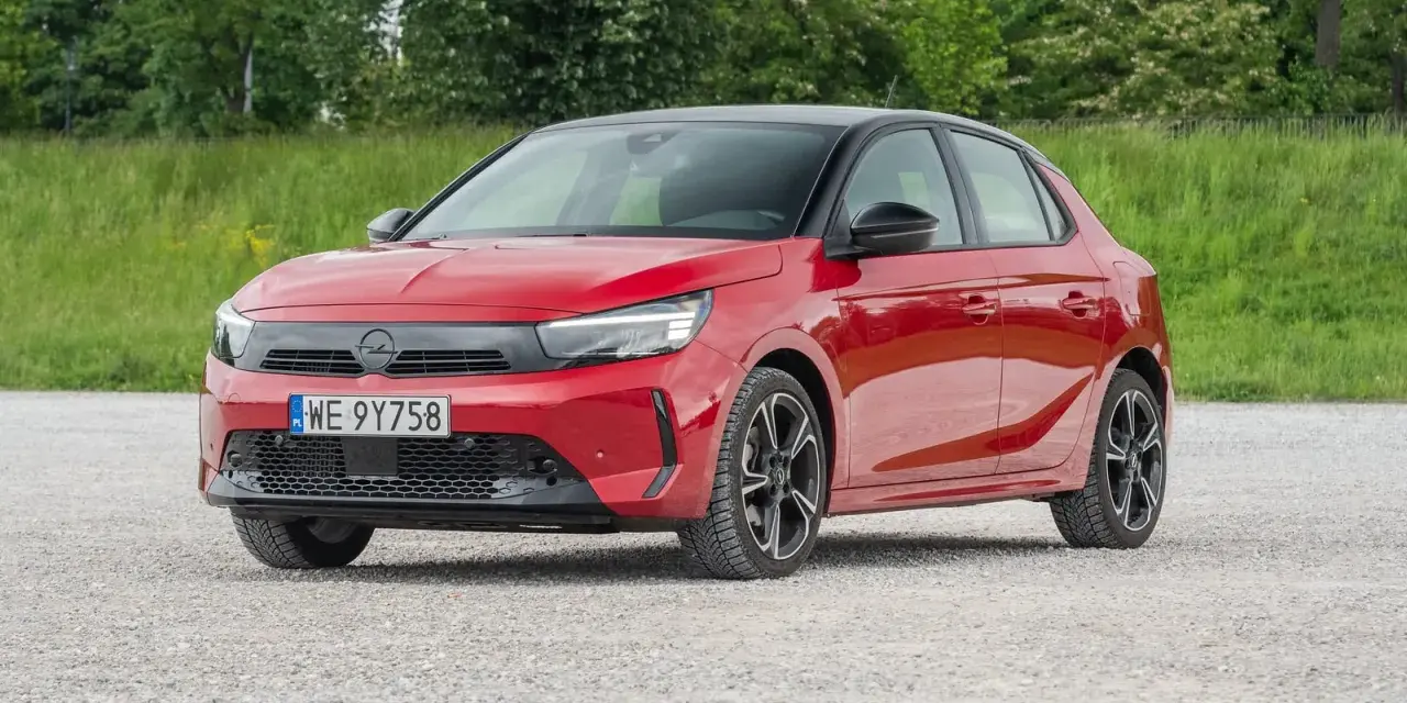 Czerwony Opel Corsa E na tle zieleni. Zbliżenie na przód auta, gdzie widoczne są światła LED i tablica rejestracyjna. Poznaj opel corsa e wymiary.