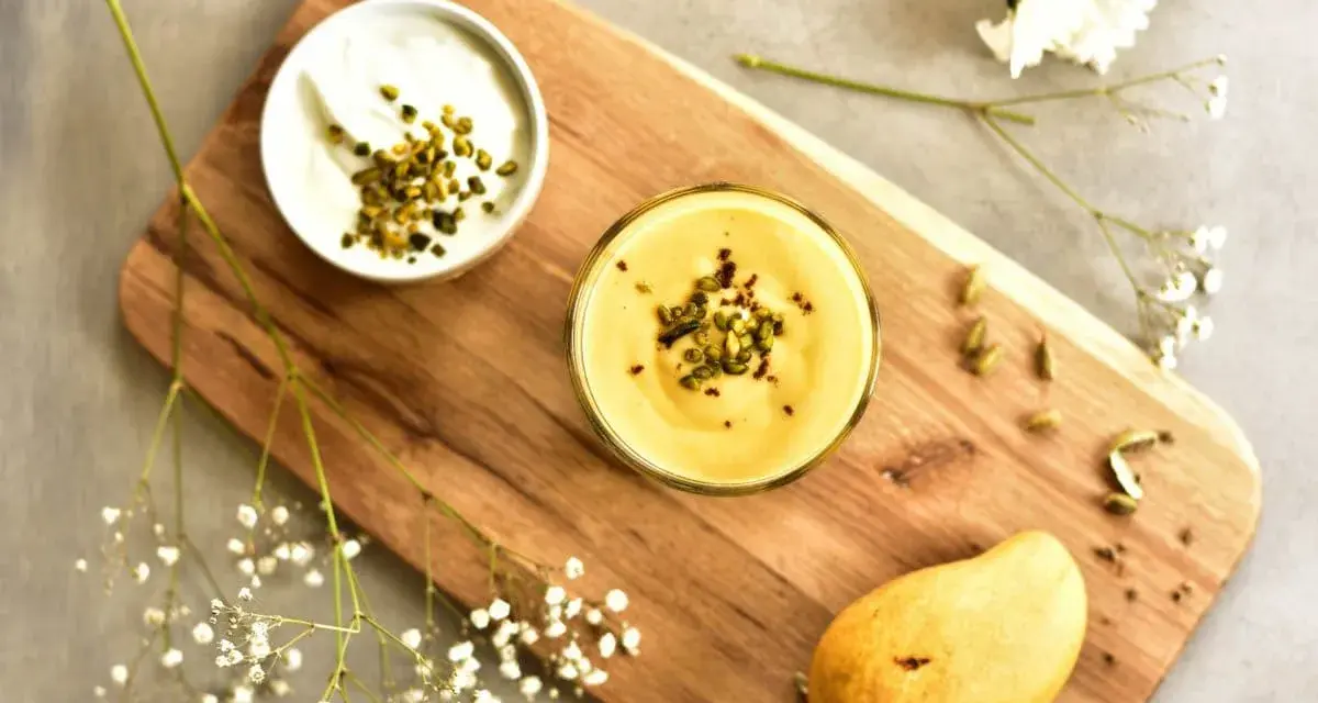 Aksamitny mango lassi w szklance, posypany pistacjami. Obok miseczka jogurtu, dojrzałe mango i kardamon.
