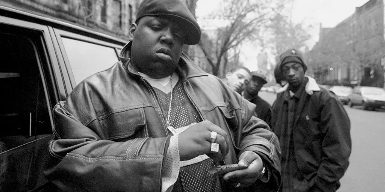 The notorious B.I.G. jak zginął. Raper w skórzanej kurtce i czapce, z towarzyszami, przed samochodem.