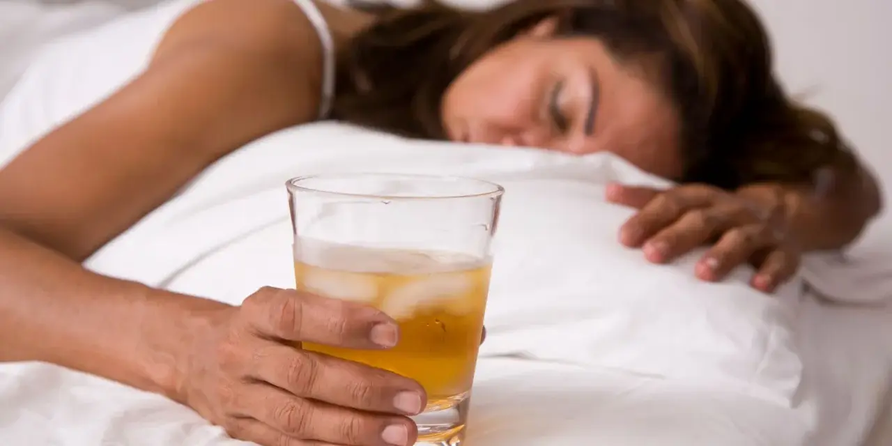Kobieta w łóżku z drinkiem, nieświadoma. Objawy po zażyciu pigulka gwaltu mogą być różne.