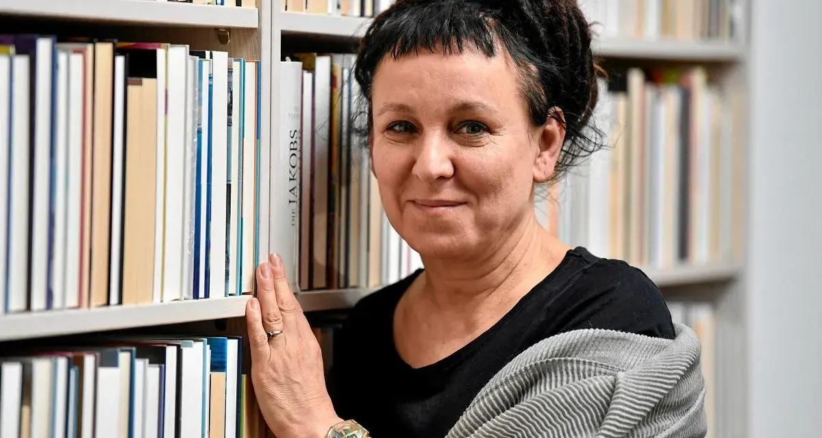 Olga Tokarczuk, w swoim wieku, otoczona książkami, z dłonią na grzbietach.