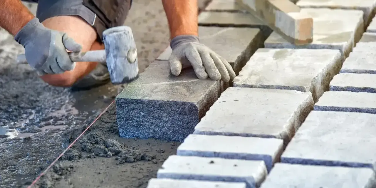 Ręce w rękawiczkach układają kamienne kostki na podsypce. Widać młotek i sznur geodezyjny. Prawidłowe suchy beton proporcje to klucz do trwałej nawierzchni.