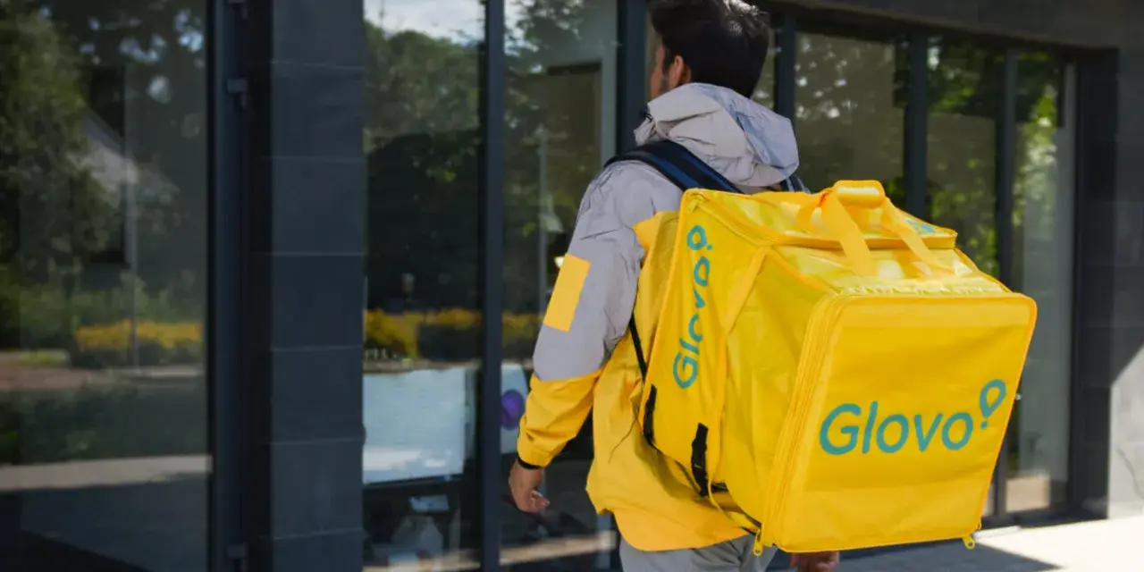 Kurier Glovo pieszo idzie z żółtą torbą na plecach.