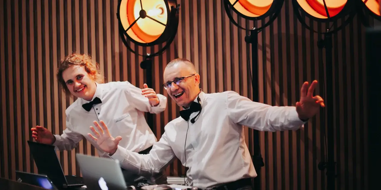 Dwóch DJ-ów w białych koszulach i czarnych muszkach, z uśmiechem na twarzy, przygotowuje się do imprezy. Zastanawiasz się, ile kosztuje DJ na wesele?