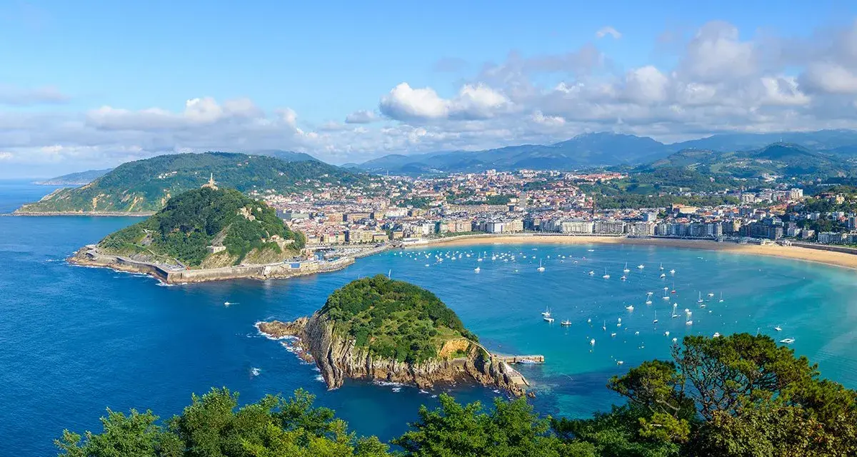 Widok na zatokę La Concha w Donostia San Sebastian, z zielonymi wzgórzami, miastem i licznymi łodziami na turkusowej wodzie.