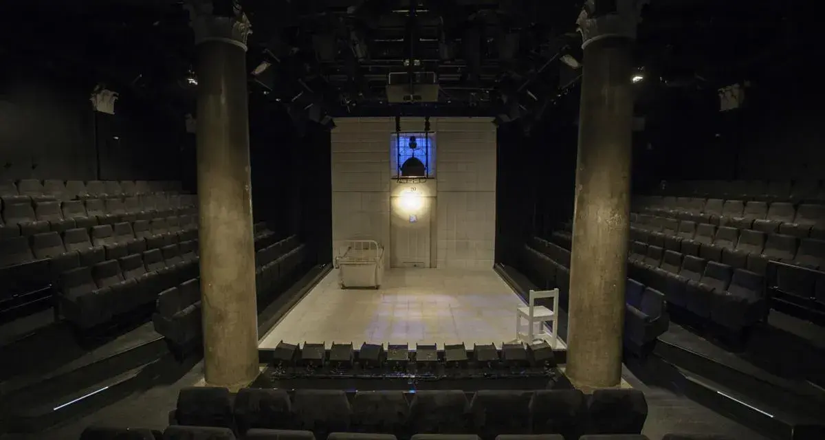 Pusta scena w **teatrze stu** miejsc, z rzędami siedzeń po bokach. Na scenie łóżko, białe krzesło i reflektory.
