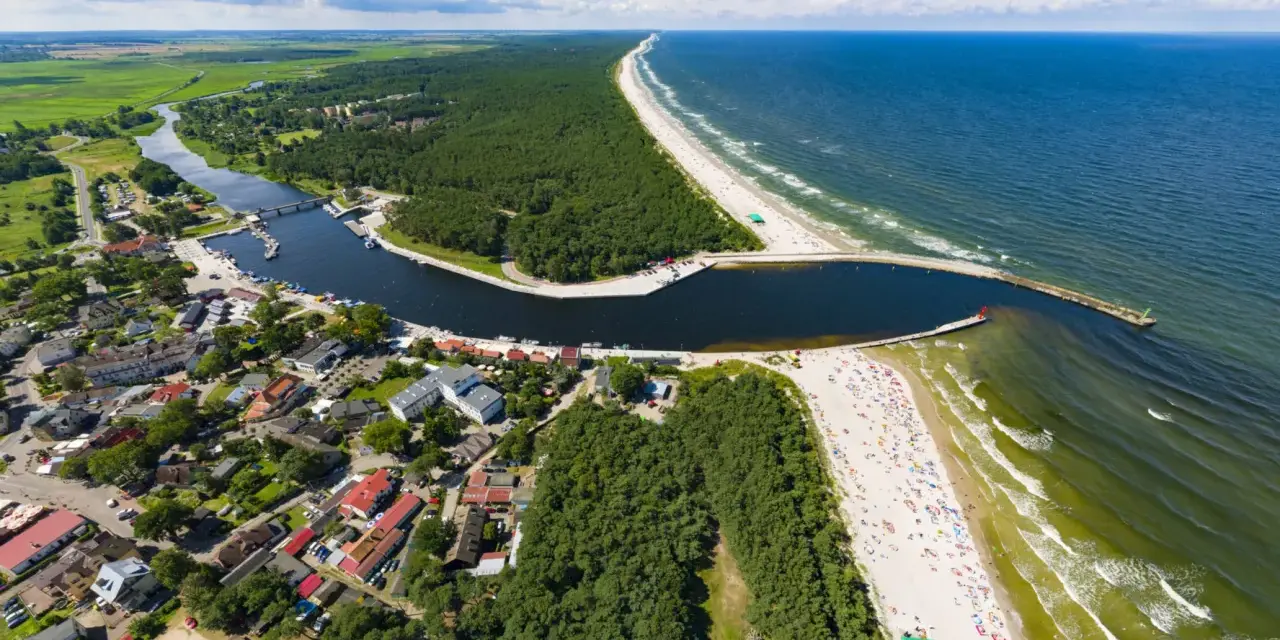 Widok z lotu ptaka na miasteczko nad zatoką, las i plażę. Na falochronie widać czerwoną latarnię morską kikut.