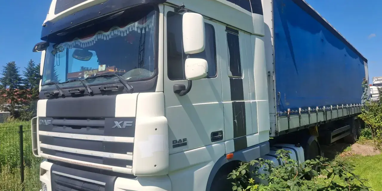 Ciężarówka DAF XF 105 z niebieską plandeką stoi na trawie, częściowo zasłonięta przez krzaki.