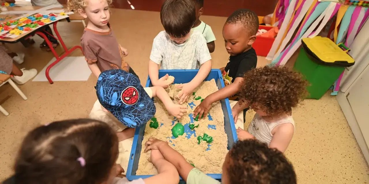 Des enfants s'amusent dans un bac à sable au centre de loisirs. Un petit garçon porte une casquette Spiderman. Ils ont tous environ 2 ans.