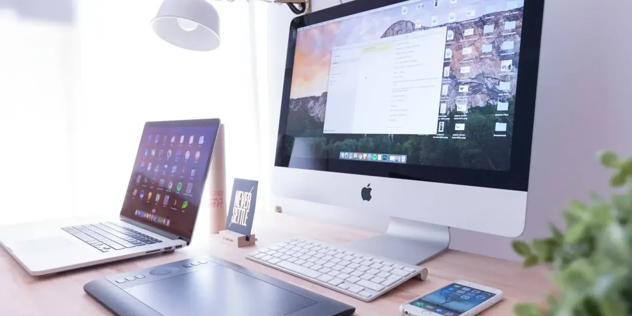 Biurko z iMac, laptopem, tabletem graficznym i telefonem. Na ekranie komputera widać okno programu Adobe Dreamweaver, gotowe do pracy nad stroną.