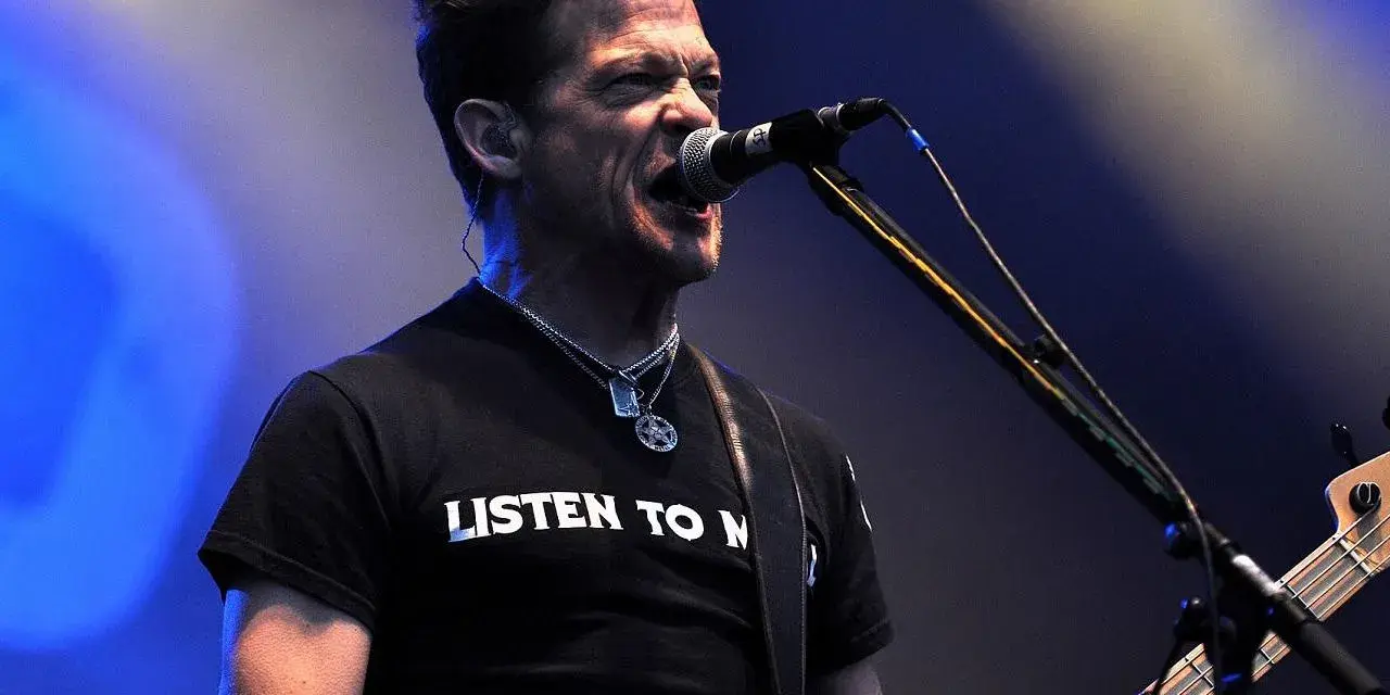 Jason Newsted z zespołu, w czarnej koszulce z napisem "LISTEN TO N", śpiewa do mikrofonu, grając na basie.