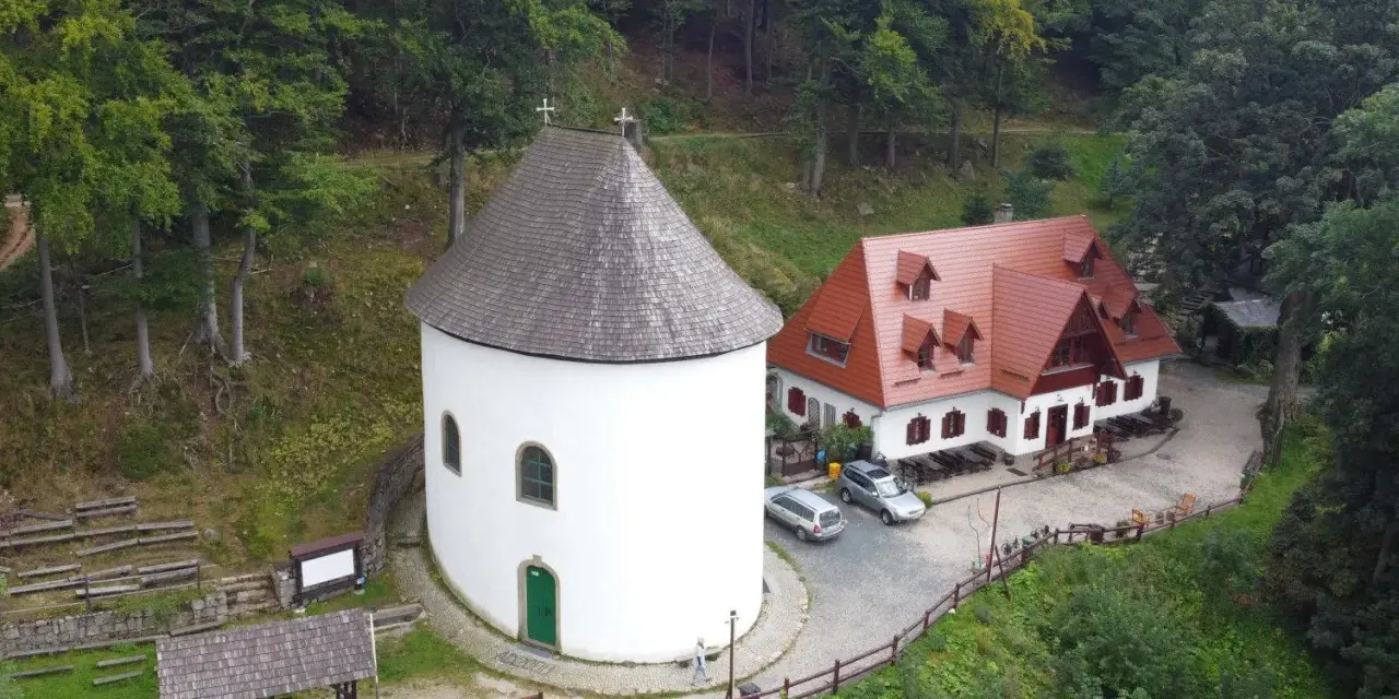 Miłków atrakcje: biała wieża z krzyżem i czerwony budynek z dachówką, otoczone zielonym lasem.