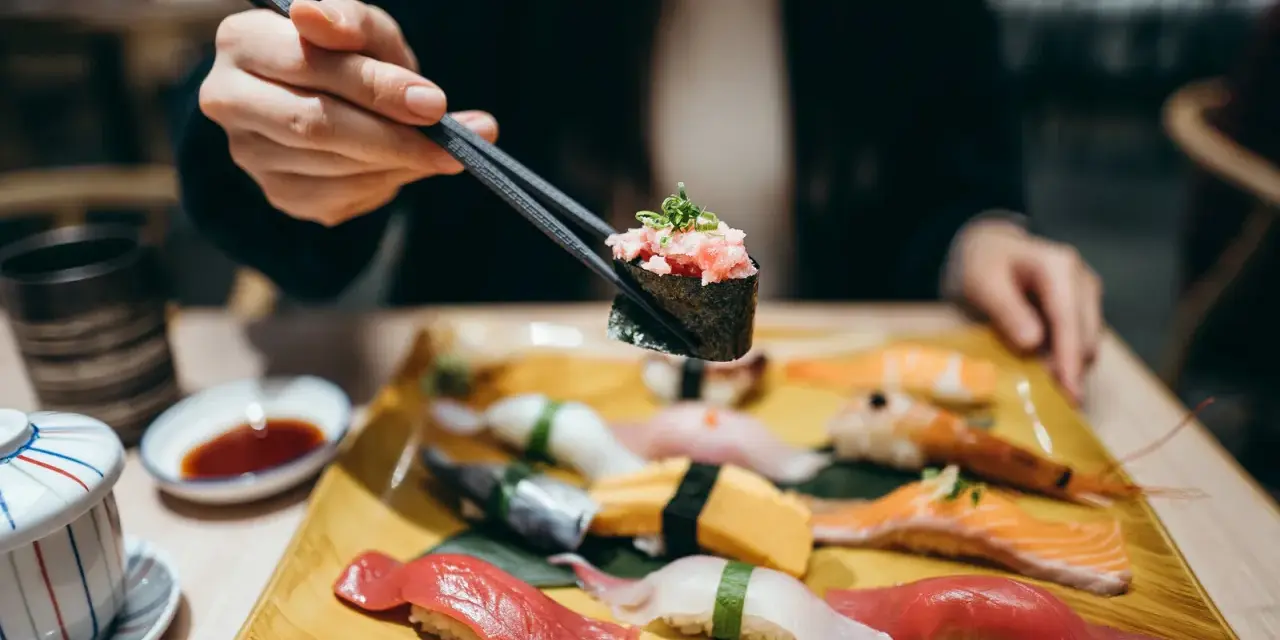 Eine Hand hält mit Stäbchen ein Stück Sushi mit gehacktem Thunfisch. Auf dem Teller liegen verschiedene Sushi-Rollen und Nigiri, bereit für den Genuss in Berlin.