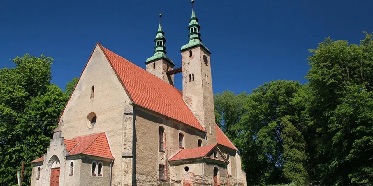 Stary kościół w stolcu z dwiema wieżami i zielonymi dachami, otoczony bujną zielenią drzew pod błękitnym niebem.