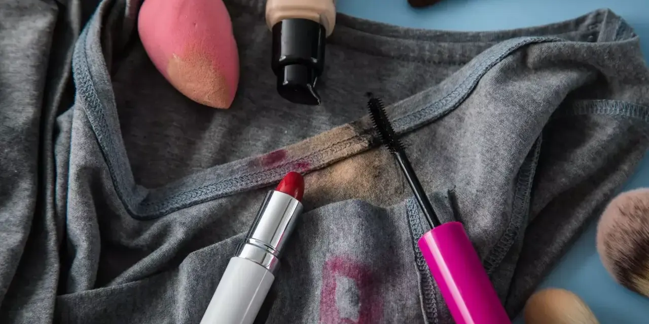 Lippenstiftflecken entfernen: Roter Lippenstift, Mascara, Foundation und Make-up-Schwamm auf grauer Kleidung.