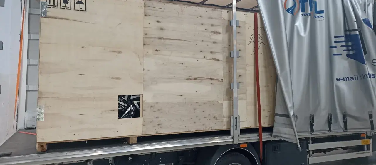 Drewniana skrzynia z widocznym fragmentem felgi, gotowa do transportu. Widać logo firmy transportowej i oznaczenia towaru.