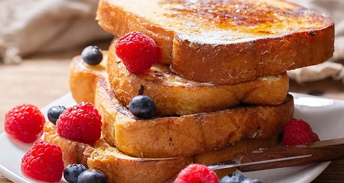 Una pila de tostadas francesas doradas, decoradas con frambuesas y arándanos, perfectas para aprender cómo se hacen las tostadas francesas.