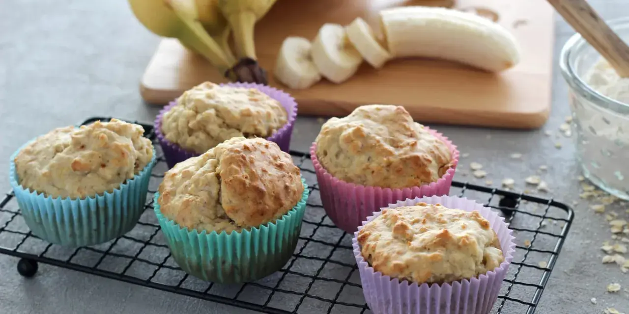 Fünf leckere Bananen Muffins in bunten Förmchen, bereit für Kinder. Bananen und Mehl sind im Hintergrund zu sehen.