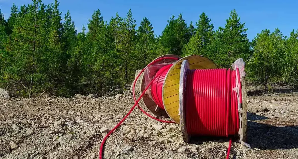 Czerwony kabel na drewnianym bębnie, przygotowany do uzbrojenia działki. W tle las i błękitne niebo.