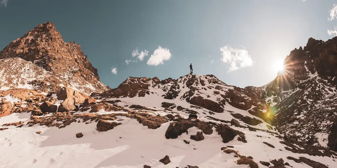 Ein Wanderer steht auf dem Gipfel des Jbel Toubkal, umgeben von schneebedeckten Felsen und einem strahlenden Sonnenschein.