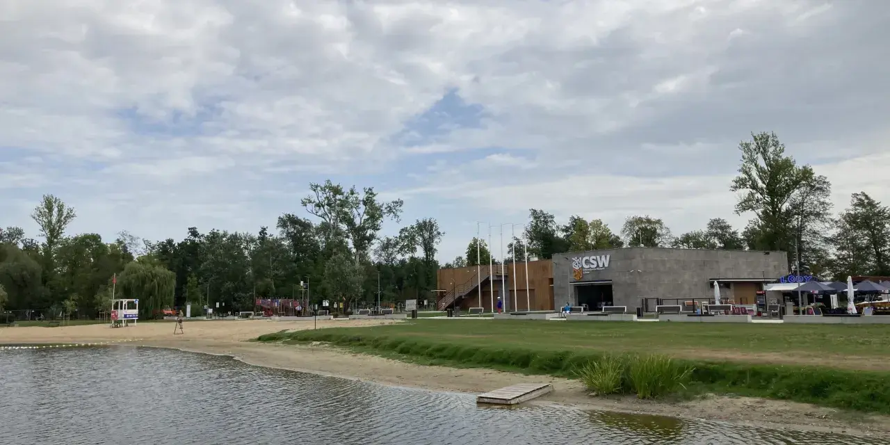 Spokojne jezioro małe żnińskie z piaszczystą plażą, budynkiem CSW i zielenią w tle.