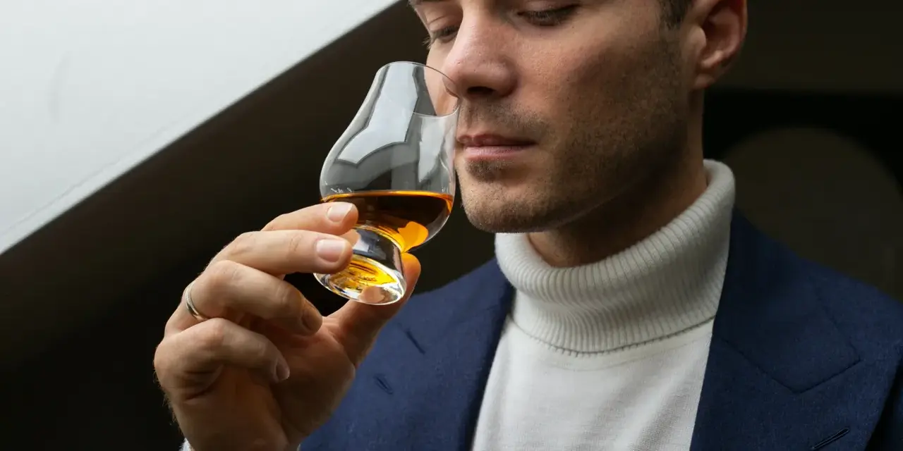 Mężczyzna w eleganckim garniturze i golfie wącha bursztynowy płyn w kieliszku. Czy to single malt?