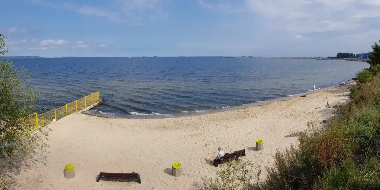 Piękna, piaszczysta plaża dla psów z łagodnym zejściem do morza. W oddali widać falochron i statki.