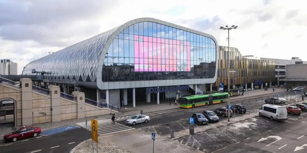 Nowoczesny Poznań Główny z charakterystyczną szklaną fasadą i ekranem. Autobusy i samochody czekają na pasażerów.