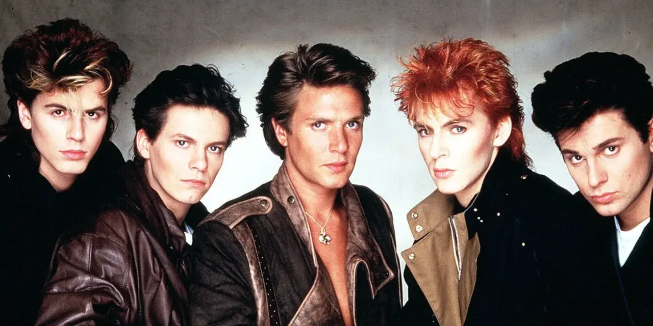 Fünf Männer mit auffälligen Frisuren und 80er-Jahre-Mode, die an ikonische Musikvideos dieser Ära erinnern.