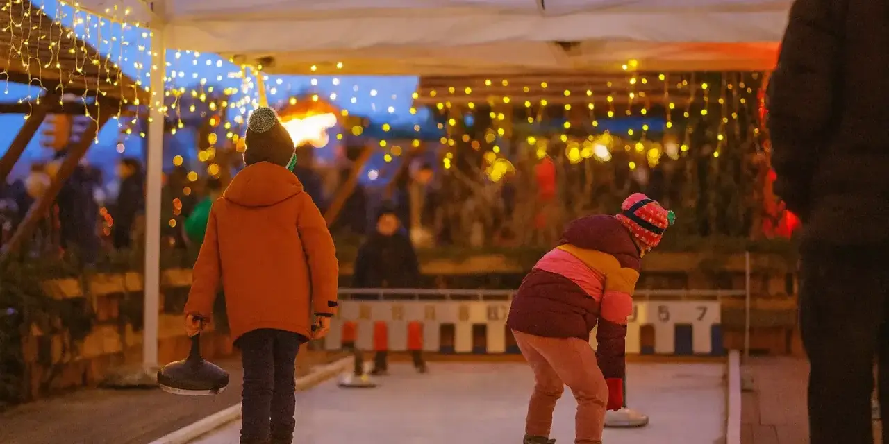Kinder spielen Eisstockschießen auf einem Weihnachtsmarkt in Berlin. Lichterketten leuchten im Abendlicht.