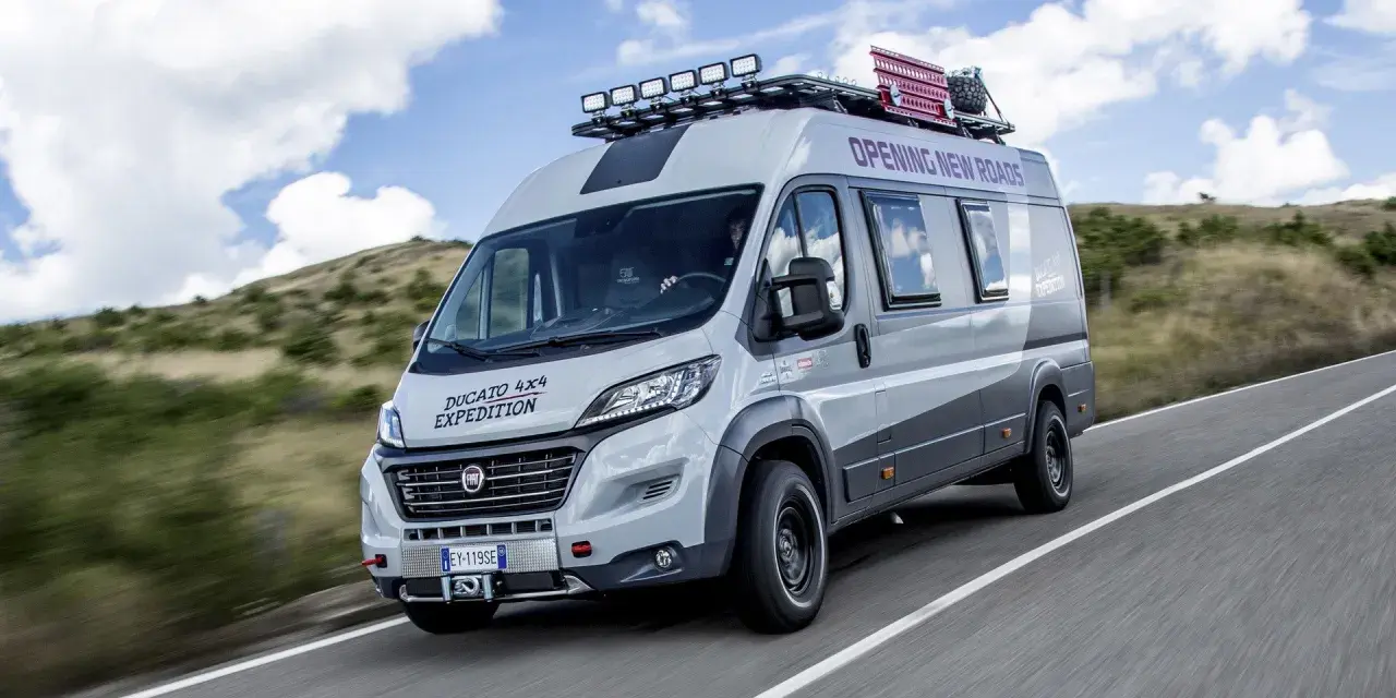 Szary kamper Ducato 4x4 Expedition z bagażnikiem dachowym i dodatkowym oświetleniem jedzie po drodze wśród wzgórz.