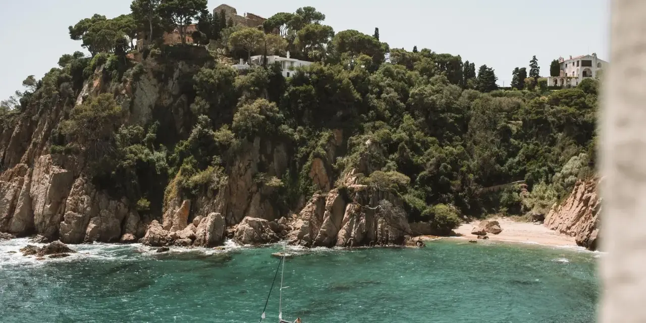 A sailboat floats in turquoise water near a rocky, tree-covered cliff with buildings. Perfect for weekend trips from Barcelona by train.