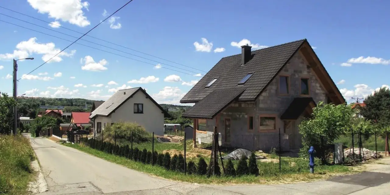 Dwa domy w budowie, jeden gotowy, na tle błękitnego nieba z chmurami. Widać drogę i zieleń. Różne kategorie gleby widoczne na poboczu.