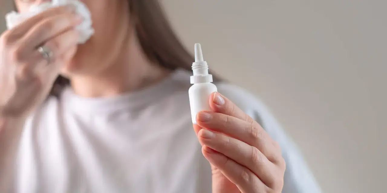 Kobieta z chusteczką przy nosie trzyma buteleczkę z kroplami do nosa, przygotowując się do plukania zatok.