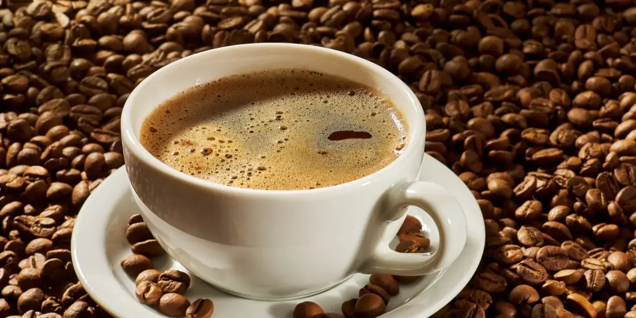 A white cup of coffee sits on a saucer surrounded by roasted coffee beans. Czy kawa rozpuszczalna podnosi ciśnienie?