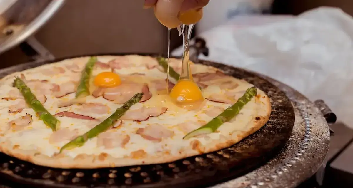 Ręka wylewa jajko na pizzę z boczkiem i szparagami. Pyszna pizza z jajkiem w przygotowaniu.