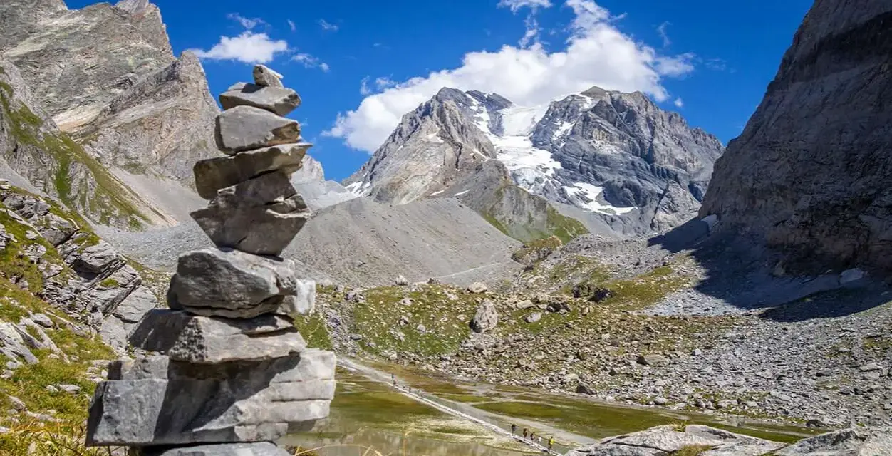 Un cairn, une pile de pierres, marque le chemin dans un paysage de montagne spectaculaire, avec un glacier au loin.