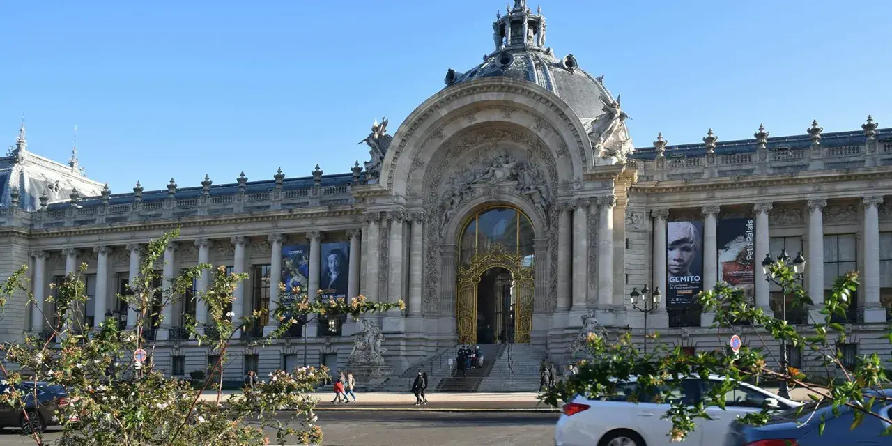 El Petit Palais, un edificio majestuoso con una cúpula y una entrada dorada, exhibe carteles de arte bajo un cielo azul.