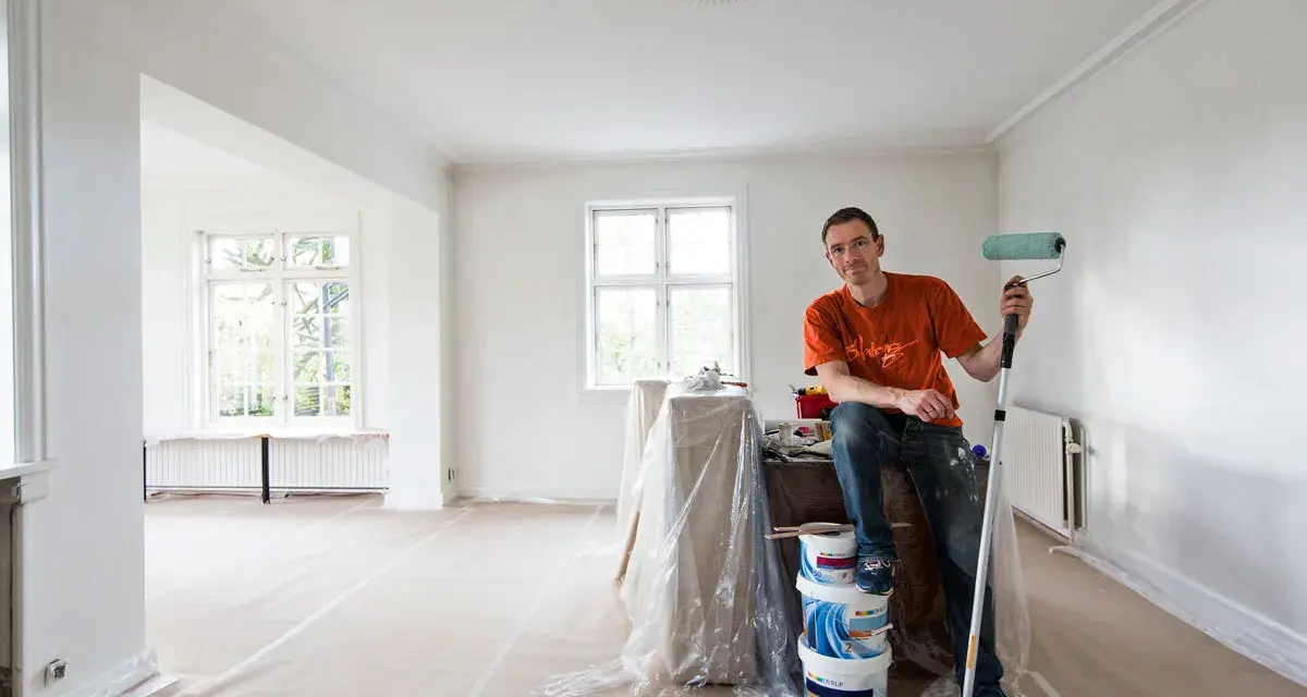 Man med färgroller och hinkar redo att måla bakom elementet i ett rum under renovering.