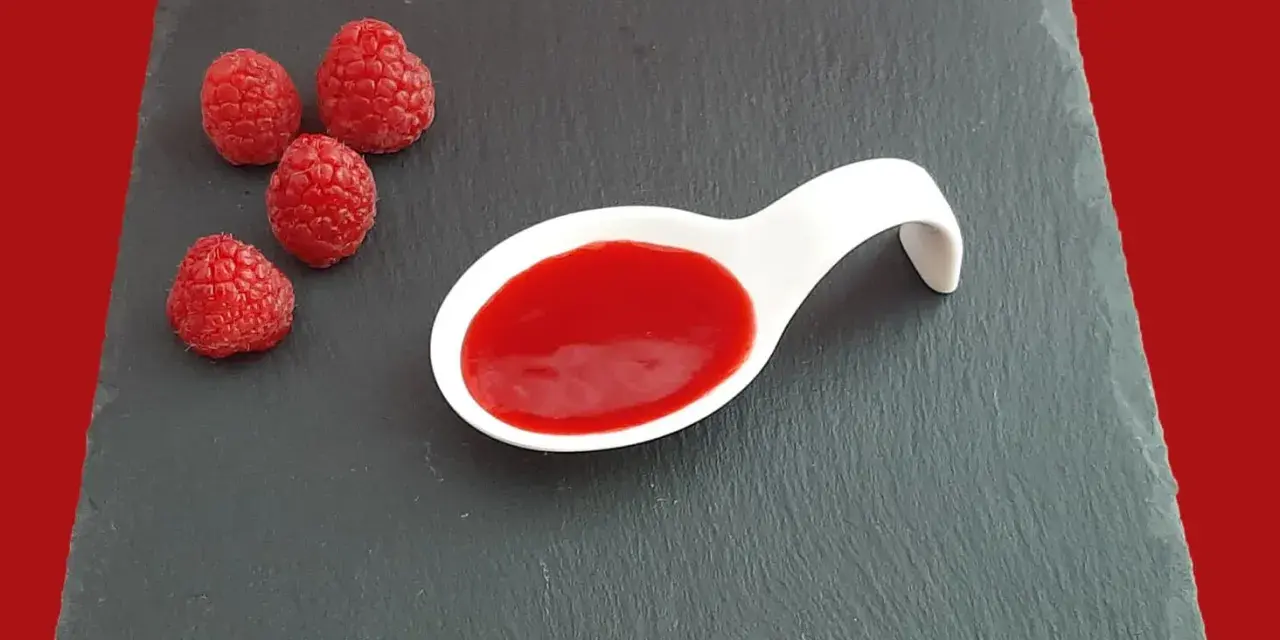 Quatre framboises fraîches à côté d'une cuillère blanche remplie de coulis de framboise, sur une ardoise.